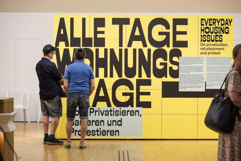 Ausstellungsansicht von einer gelben Wand auf der "Alle Tage Wohnungsfrage" in schwarzer Schrift steht, davor Besucher, die den Text lesen